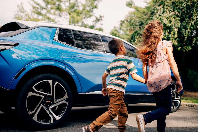 Two children holding hands walking by a blue Chevy vehicle on a sunny day